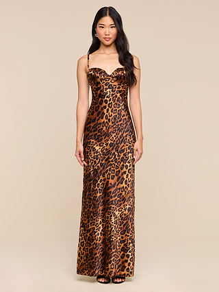 A woman stands facing forward, wearing the Sayla Silk Gown—a long, sleeveless leopard print dress with thin straps and underwire cups—paired with black high-heeled shoes. The background is plain beige.