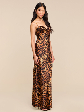 A woman with long black hair wears the Sayla Silk Gown—a sleeveless, floor-length silk dress in leopard print with spaghetti straps—paired with black high heels, posing against a plain beige background.