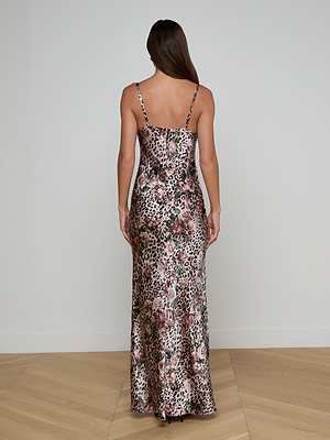 A woman with long brown hair faces away, wearing the Sayla Silk Gown—a floor-length rose cheetah print dress with thin spaghetti straps. She stands on a light wooden floor against a plain white wall.