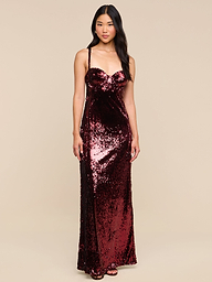 A woman stands against a plain white background, wearing the Sayla Sequin Gown—a long, sleeveless gown in dark red sequins that radiates vintage glamour and pairs elegantly with her black heels. She has long, dark hair and a relaxed, confident expression.