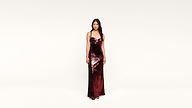 A woman stands wearing a shiny, dark red sequin gown with thin straps against a plain, light background.