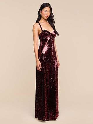 Dressed in the Sayla Sequin Gown—a floor-length, burgundy creation adorned with shimmering sequins and delicate thin straps—a woman channels vintage glamour as she poses against a plain beige background.