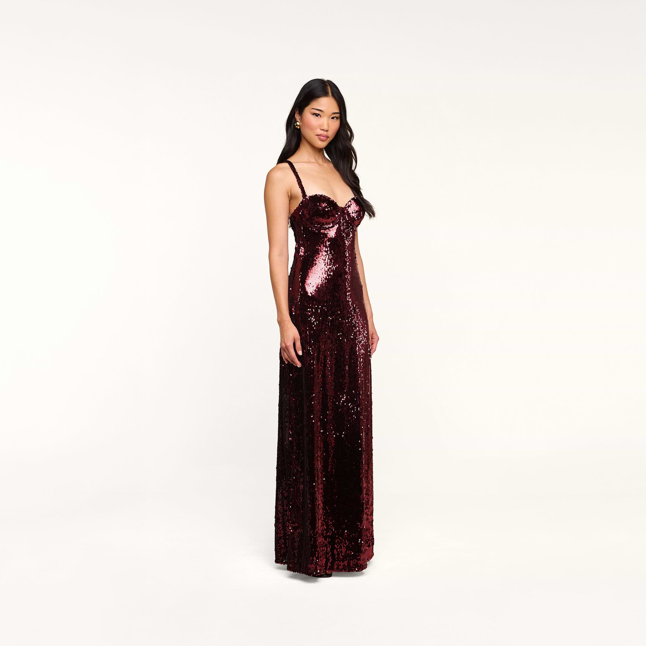 The woman stands facing forward against a plain white background, wearing the sleeveless, floor-length Sayla Sequin Gown in burgundy, a dress that exudes vintage glamour.