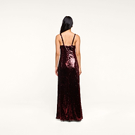 A woman with long dark hair faces away, wearing the floor-length Sayla Sequin Gown—a dark red silk dress with thin straps—standing against a plain light background for a look that captures vintage glamour.