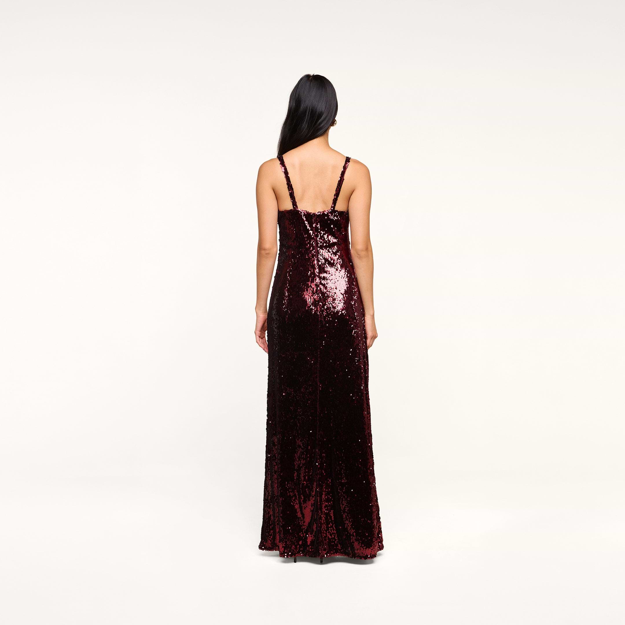 A woman with long dark hair faces away, wearing the floor-length Sayla Sequin Gown—a dark red silk dress with thin straps—standing against a plain light background for a look that captures vintage glamour.