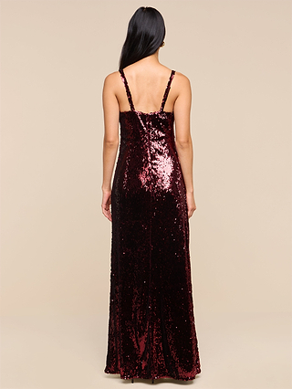 A woman stands with her back to the camera, exuding vintage glamour in the Sayla Sequin Gown—a long, dark red dress with thin straps—set against a plain beige background.