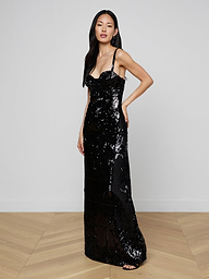 A woman poses against a plain background wearing the Sayla Sequin Gown, a long black dress adorned with sequins and delicate thin straps. Her long, straight hair complements the gown’s vintage-inspired glamour as she stands with one hand on her hip, gazing calmly at the camera.