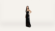 A woman stands in a long, black, sequin dress with thin straps against a plain, light background. She poses with one hand on her hip and looks forward, her dark hair worn straight.