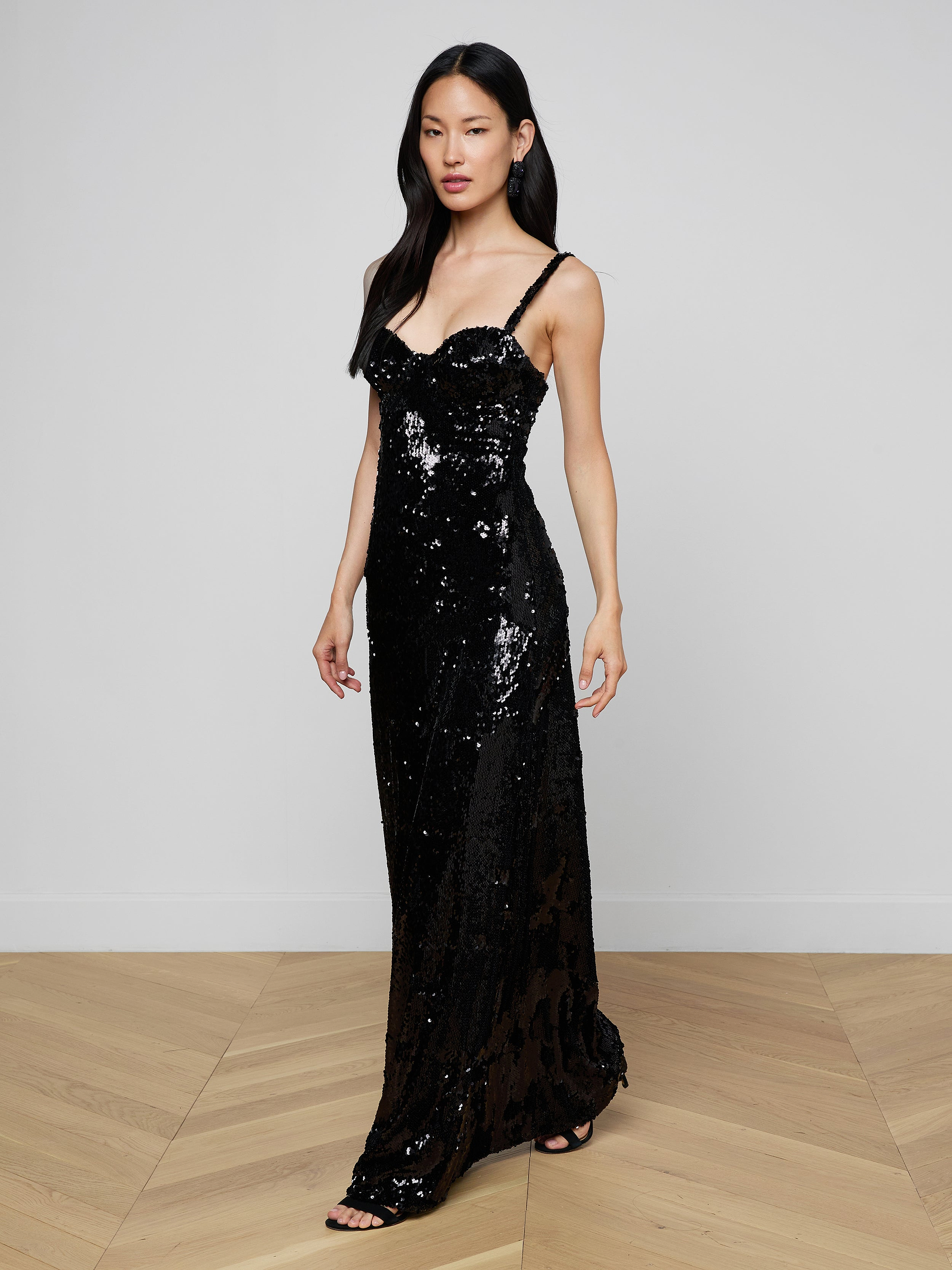A woman wearing the Sayla Sequin Gown—a long, black sequin dress with thin straps that exudes vintage glamour—stands against a plain light background.