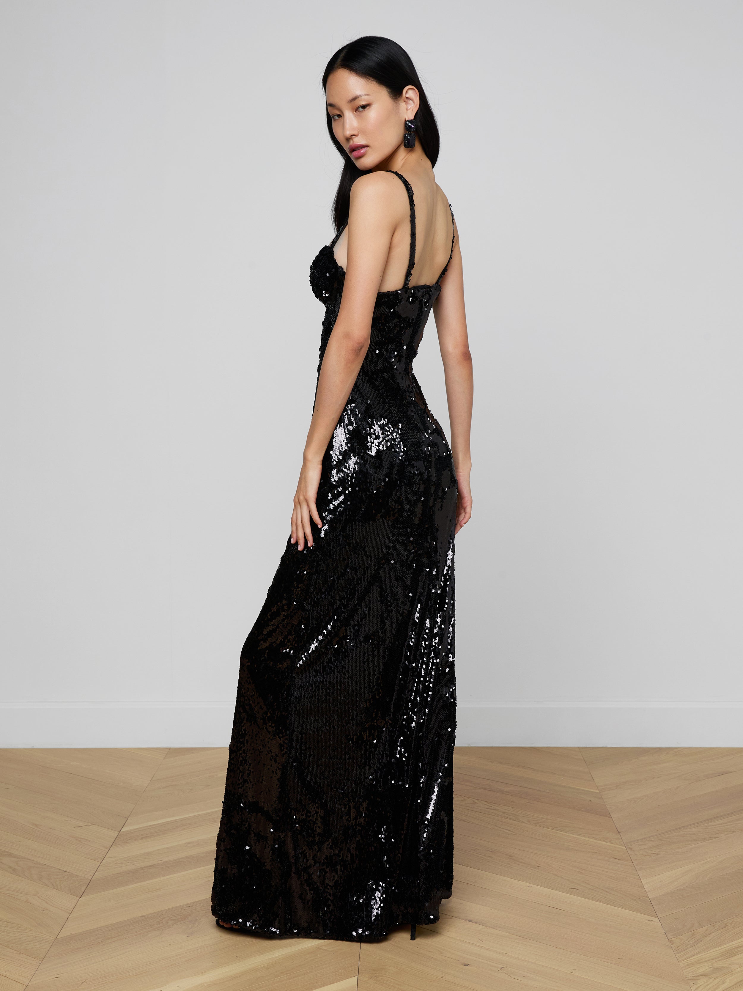 Draped in the Sayla Sequin Gown—a long, sleeveless black sequin dress—a woman stands against a plain light background, channeling vintage glamour as she gazes over her shoulder with one hand resting by her side.