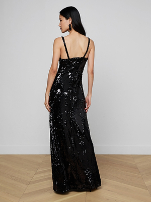 A woman with long dark hair stands facing away, wearing the Sayla Sequin Gown—black, floor-length with thin straps. She is event-ready indoors on a light wood floor against a plain white wall.