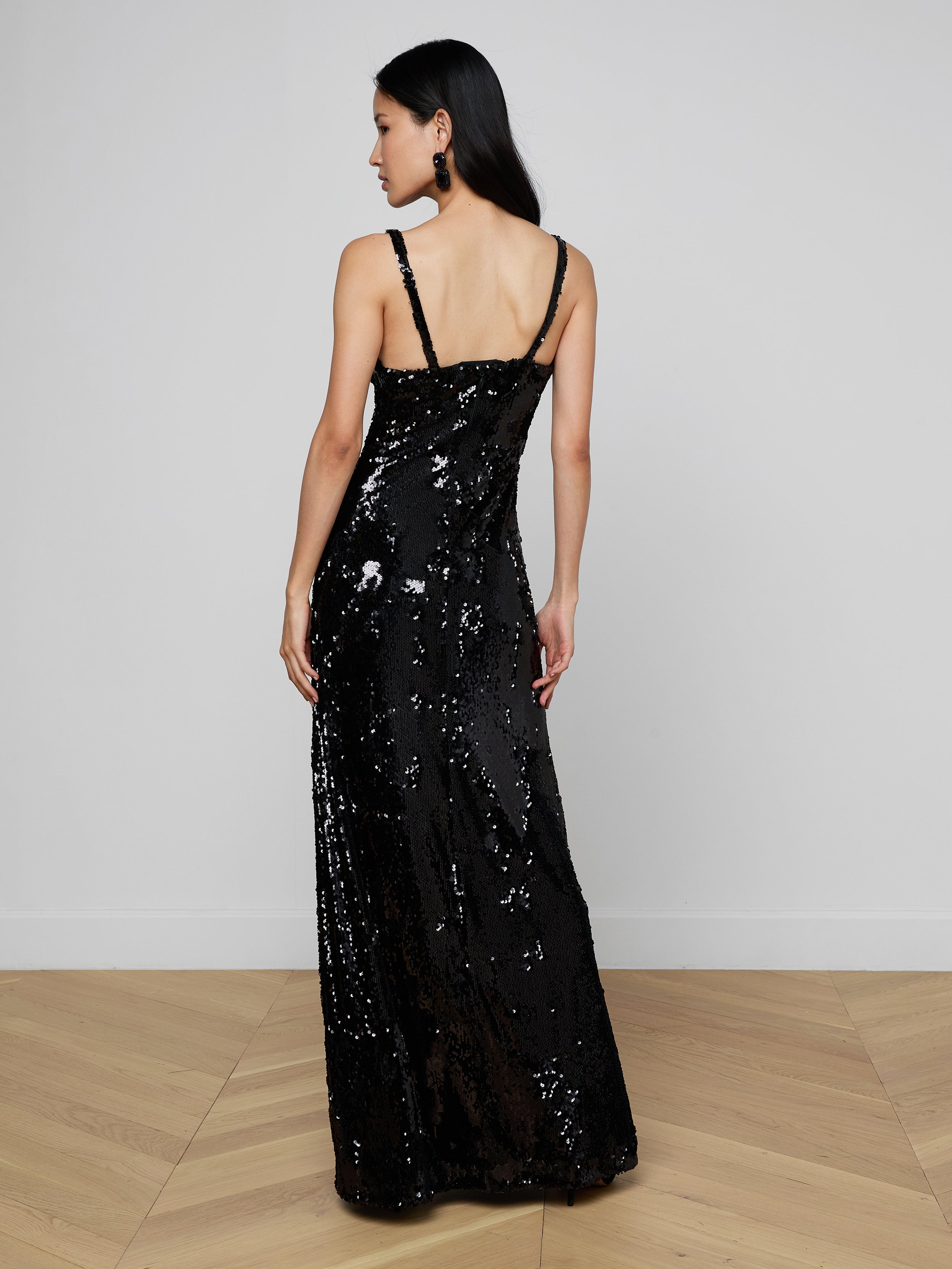 A woman stands facing away against a plain light background, showcasing the back of the Sayla Sequin Gown—a long, sleeveless black sequin dress that exudes vintage glamour.