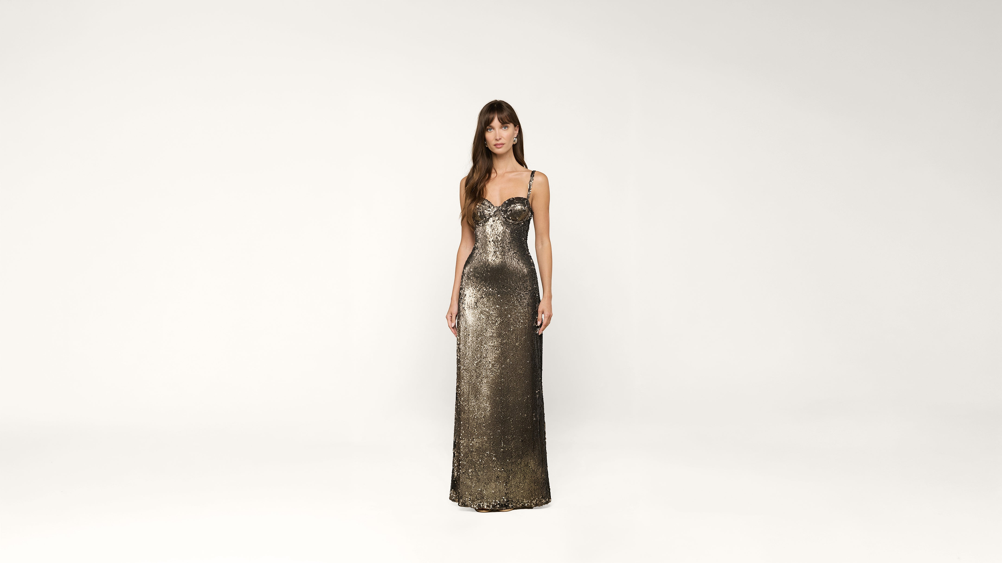 A woman in a metallic, floor-length, fitted sequin gown stands against a plain white background, looking forward with relaxed arms at her sides.