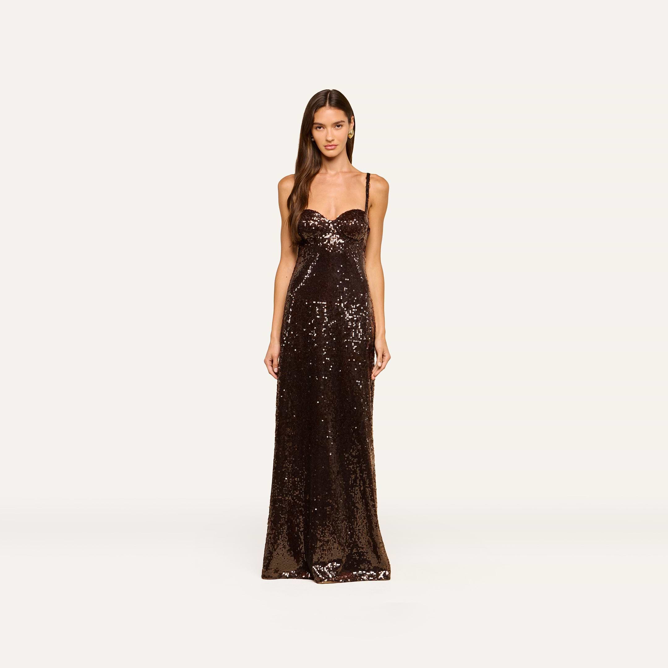 A woman stands facing forward, wearing the Sayla Sequin Gown—a floor-length, event-ready dress featuring thin straps and brown sequins—against a plain, light background.