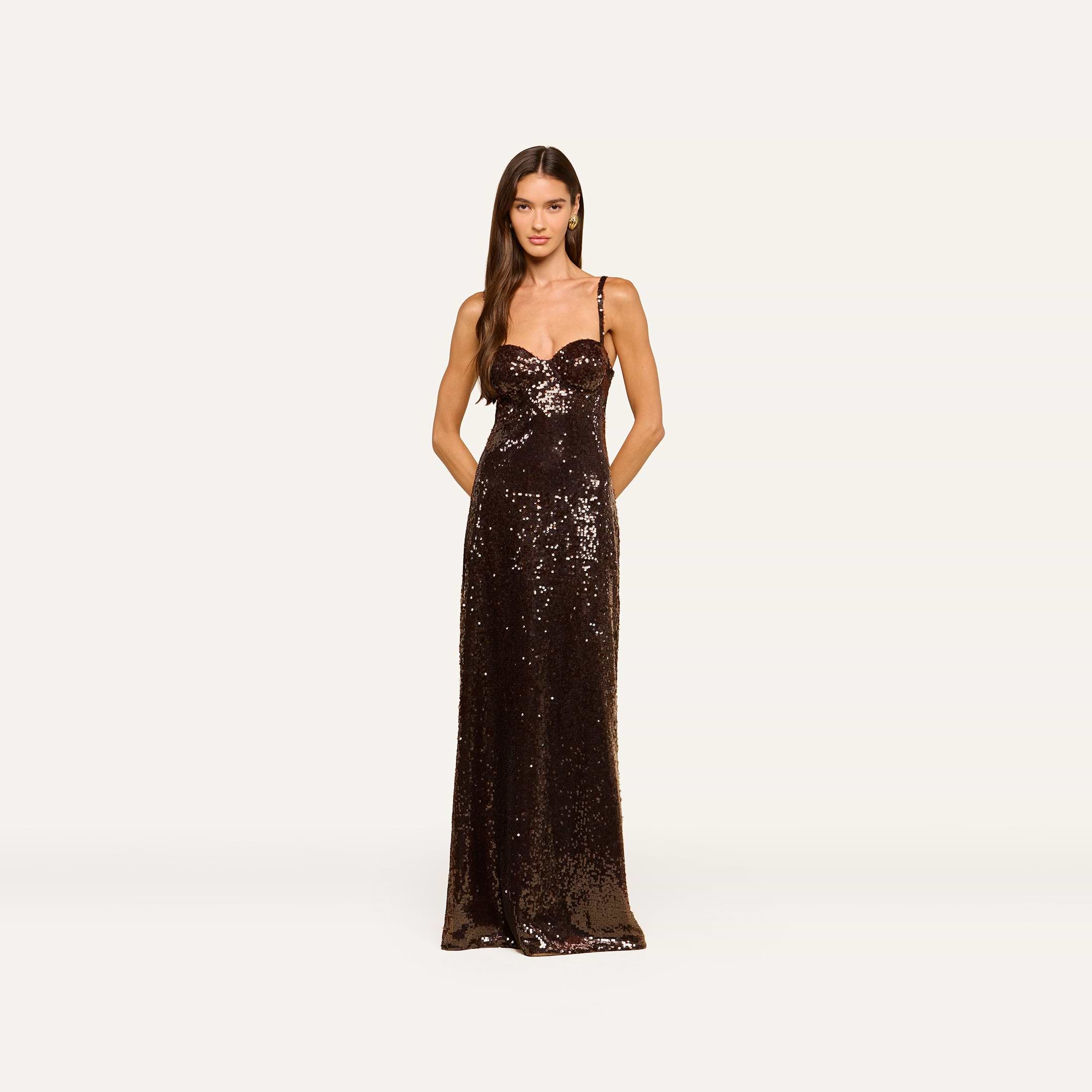 A woman stands facing forward, wearing the Sayla Sequin Gown—a long, sleeveless dress in dark brown with thin straps and pockets—against a plain white background; an event-ready gown perfect for any occasion.