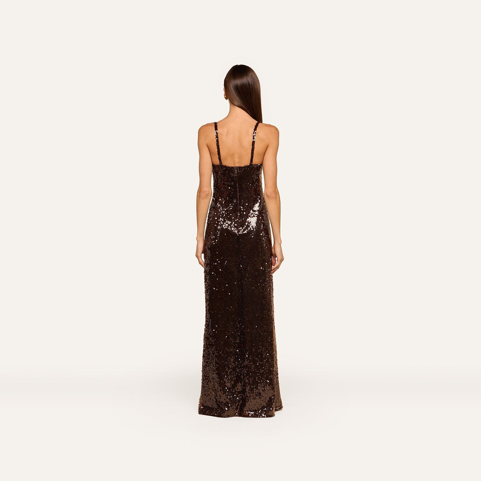 A woman with long straight hair stands facing away, wearing the Sayla Sequin Gown—a floor-length dress in dark brown sequins with thin shoulder straps—against a plain light background, creating an event-ready look perfect for stylish occasions.