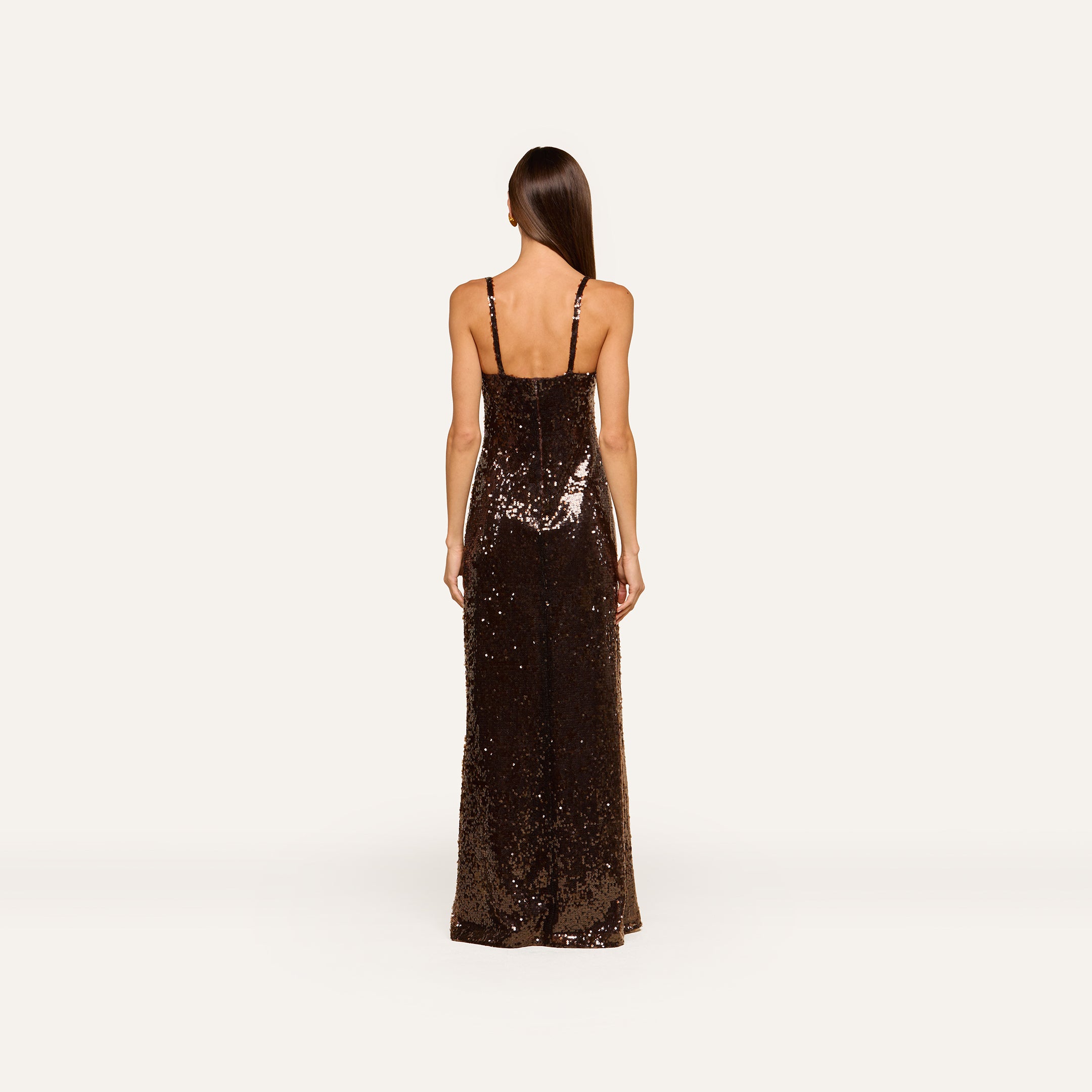 A woman with long straight hair stands facing away, wearing the Sayla Sequin Gown—a floor-length dress in dark brown sequins with thin shoulder straps—against a plain light background, creating an event-ready look perfect for stylish occasions.