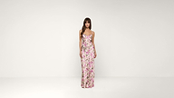 A woman stands against a plain white background, wearing a long, fitted pink floral dress with thin straps.
