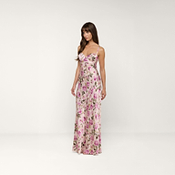 A woman wearing the Sayla Silk Gown—a long, sleeveless pink floral silk dress—stands in a plain white room, gazing slightly to the side and evoking vintage glamour.