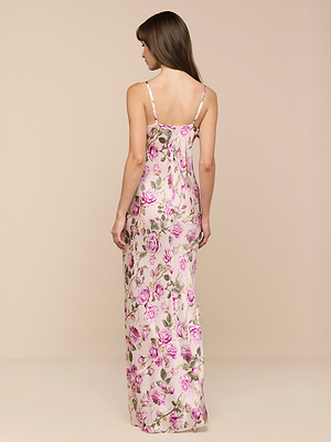 A woman with long brown hair stands with her back to the camera in a Sayla Silk Gown, a sleeveless, floor-length dress adorned with pink and green floral patterns, set against a plain beige background for vintage glamour.