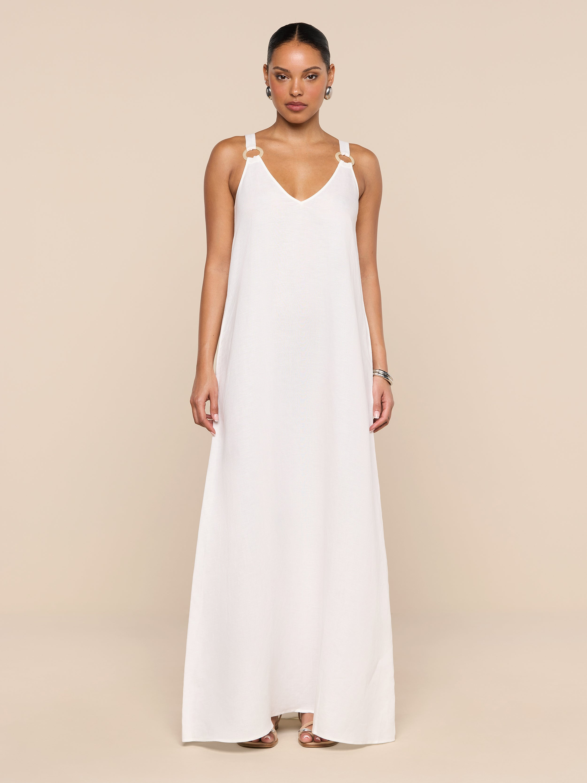 A woman stands against a plain white background wearing the Didi Maxi Dress—a long, sleeveless maxi dress featuring adjustable straps and silver embellishments on the shoulders. Her hair is pulled back, and she accessorizes with hoop earrings and sandals.