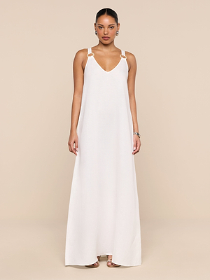 A woman stands against a beige background wearing the Didi Maxi Dress, a long white linen-blend maxi with a V-neck and gold ring accents on the adjustable straps. Her hair is pulled back, and she completes the look with earrings and bracelets.