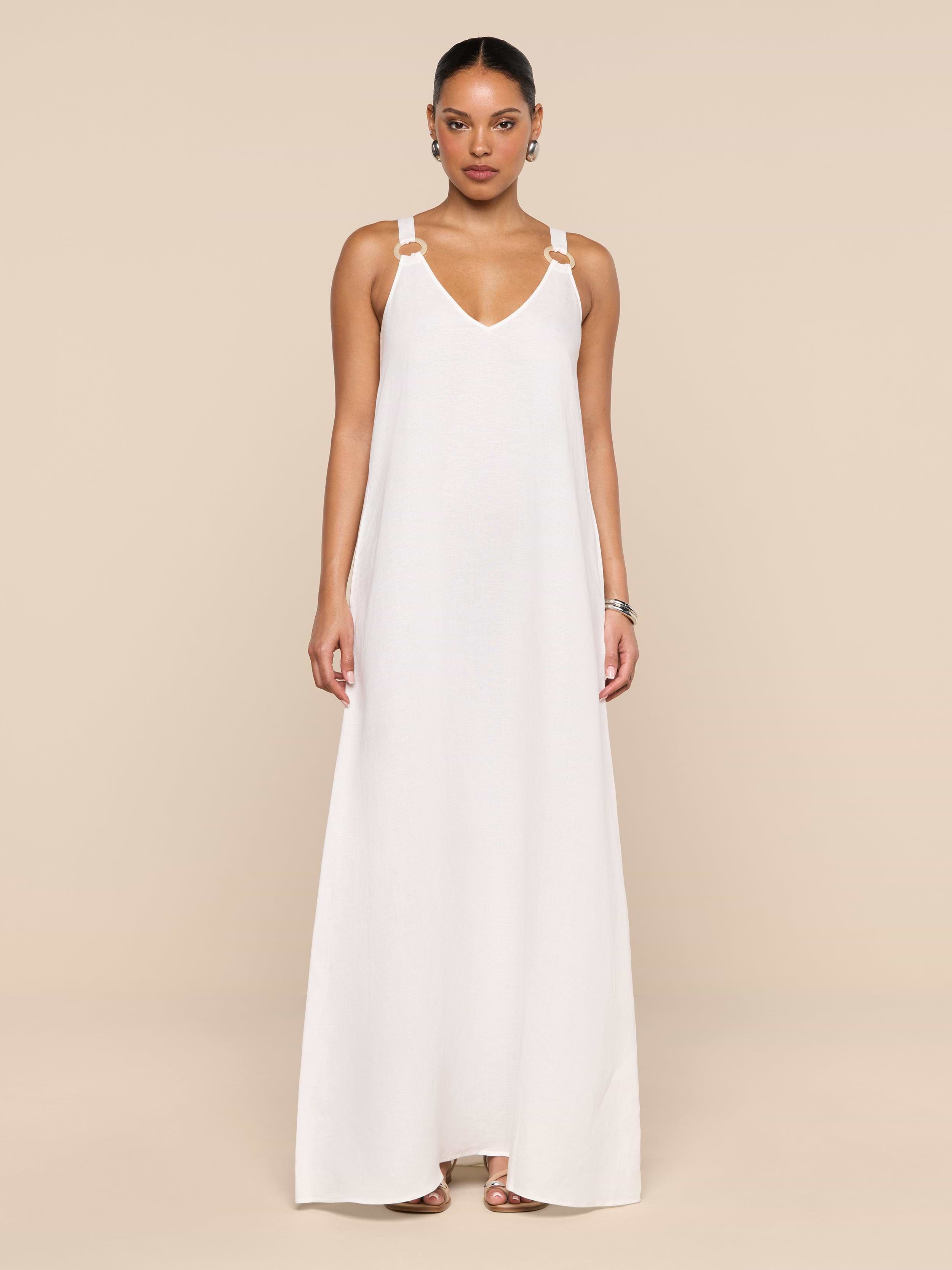 A woman stands against a plain white background wearing the Didi Maxi Dress—a long, sleeveless maxi dress featuring adjustable straps and silver embellishments on the shoulders. Her hair is pulled back, and she accessorizes with hoop earrings and sandals.