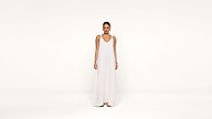 A woman stands against a plain white background, wearing a long, sleeveless white dress with thin straps and sandals. She faces forward with a neutral expression, arms relaxed at her sides.