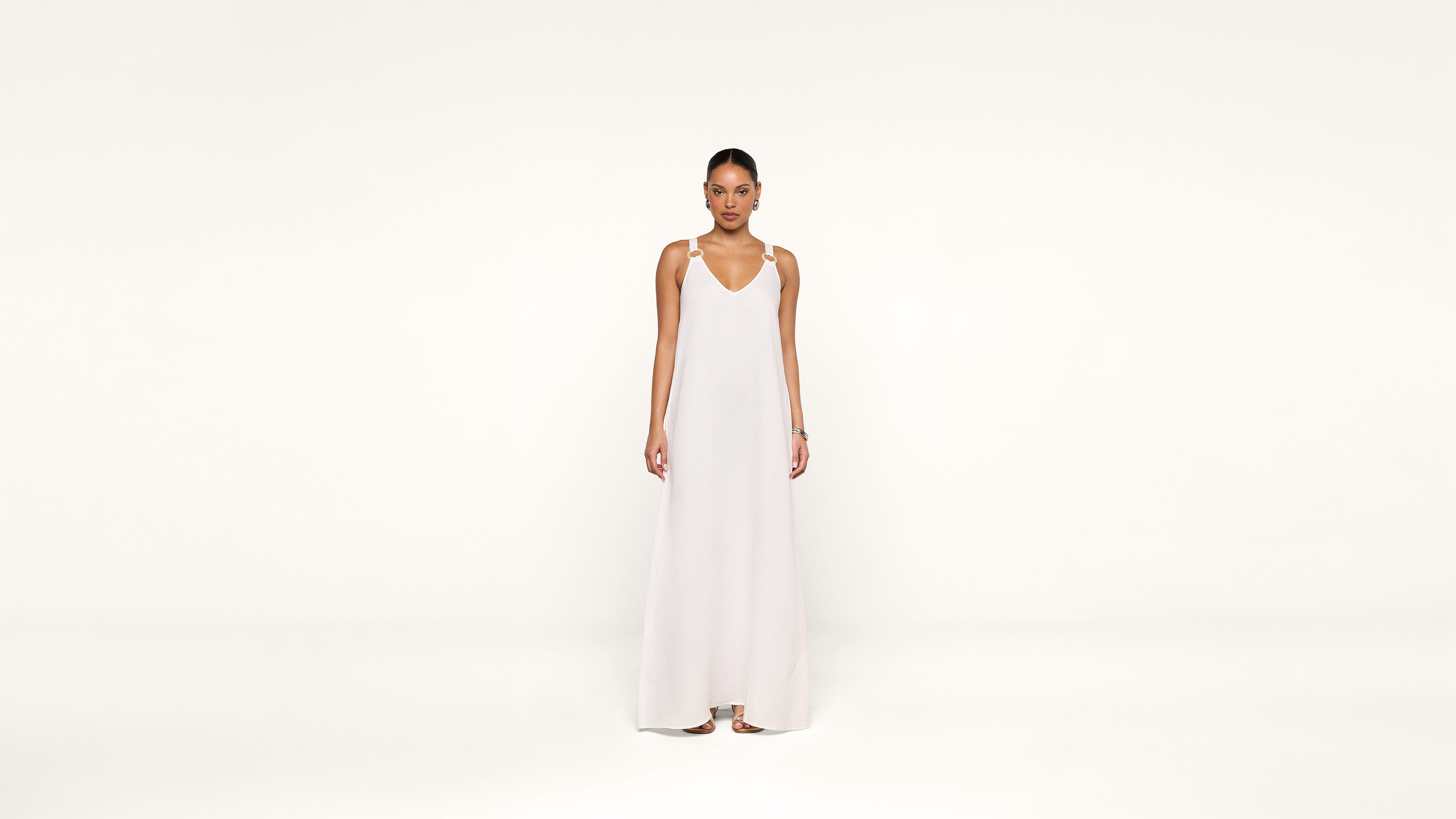 A woman stands against a plain white background, wearing a long, sleeveless white dress with thin straps and sandals. She faces forward with a neutral expression, arms relaxed at her sides.