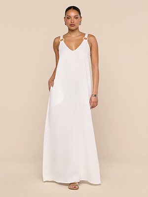 A woman stands against a beige background wearing the Didi Maxi Dress, a white linen-blend maxi with a V-neckline, gold ring accents, and adjustable straps. Her hair is pulled back, and she completes the look with earrings, bracelets, and sandals.