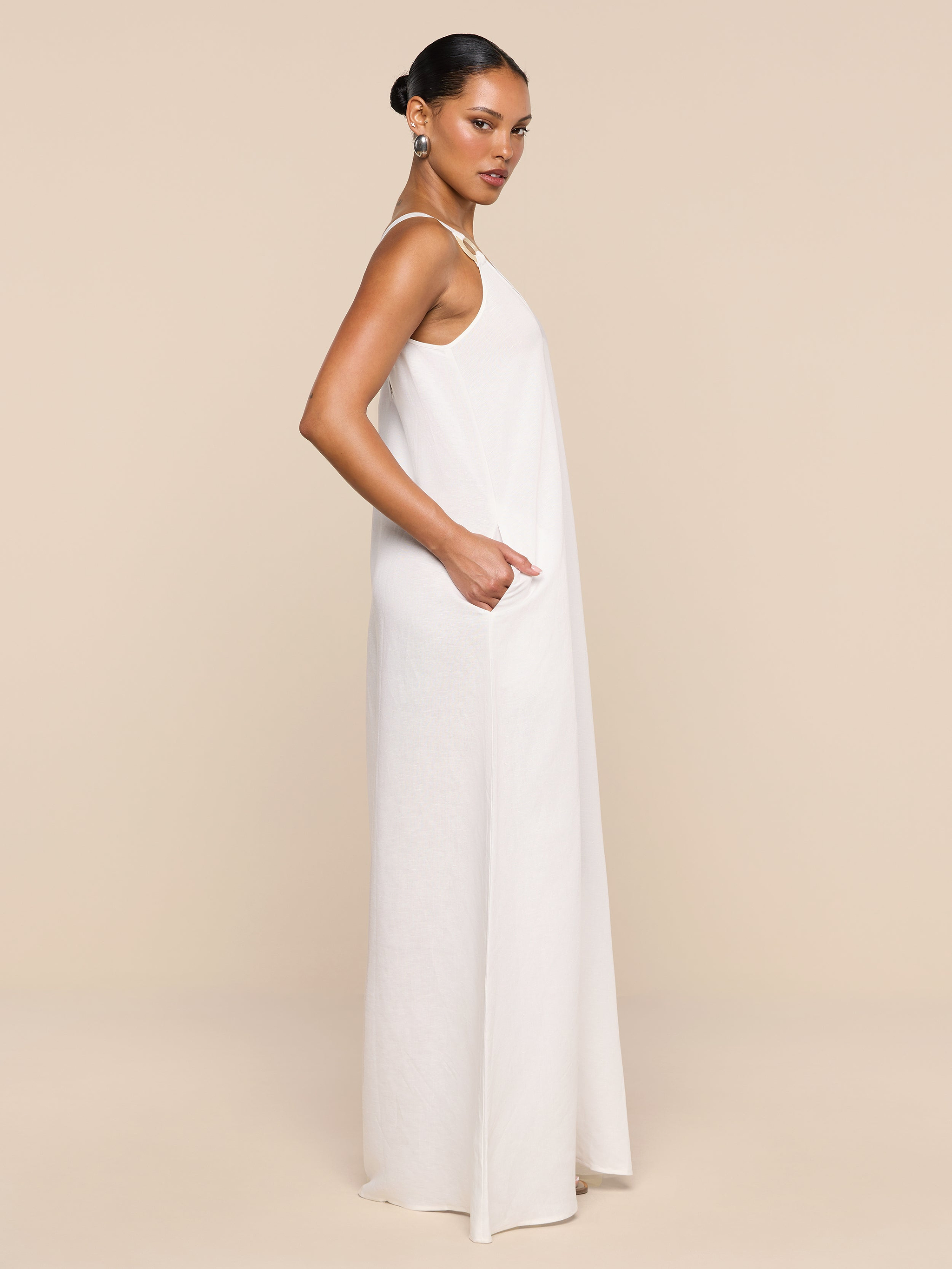 A woman stands sideways with her hands in the pockets, wearing the sleeveless, floor-length Didi Maxi Dress crafted from a linen blend. The background is plain white.