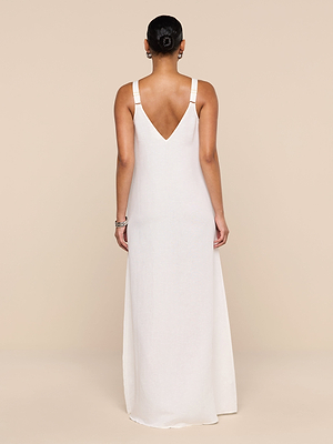 A person stands facing away, wearing the Didi Maxi Dress—a long, sleeveless white linen-blend maxi with a deep V-shaped back. Their hair is neatly pulled back, bracelets on one wrist. The background is solid beige.