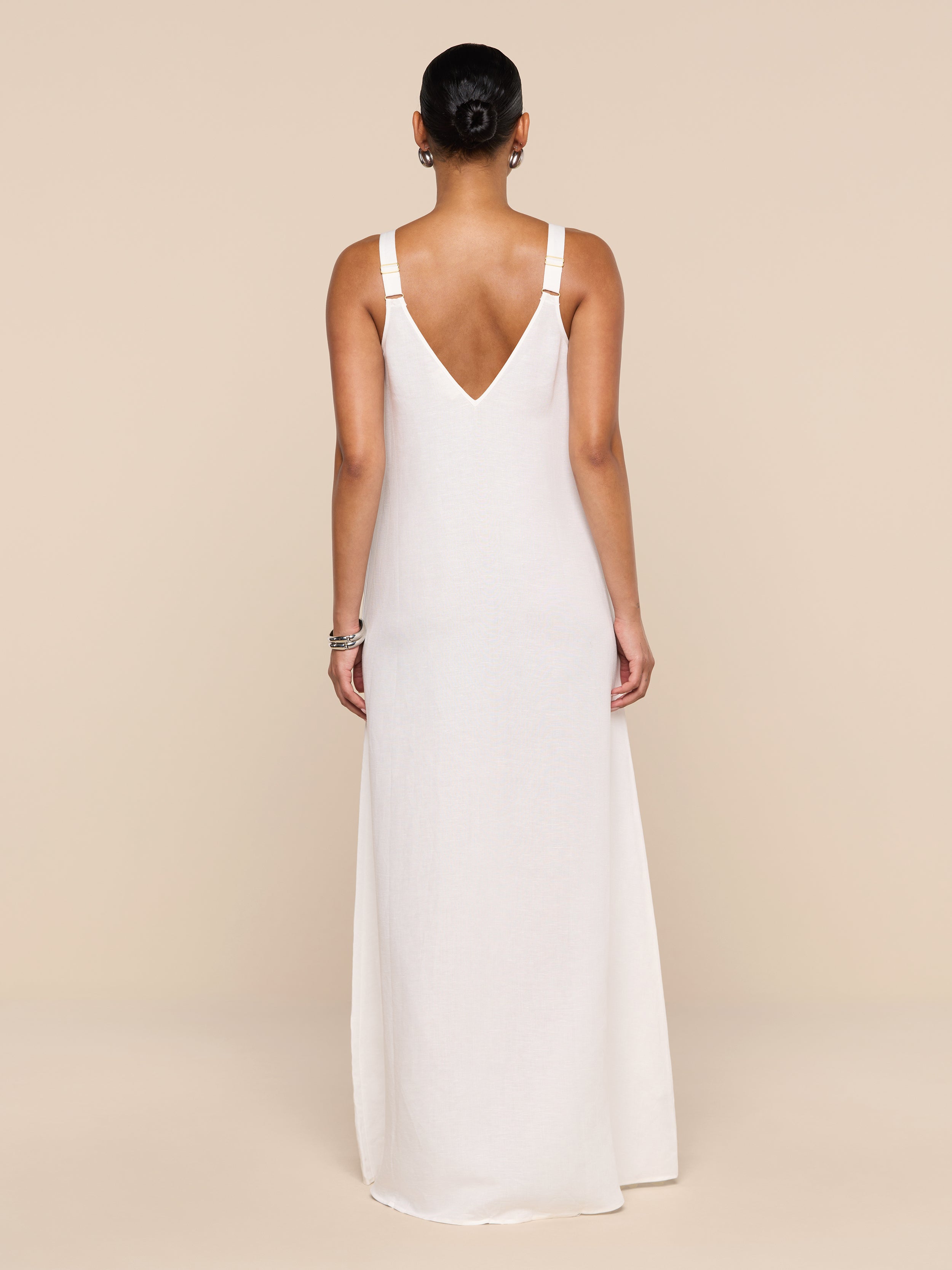 A woman stands facing away against a plain white background, wearing the Didi Maxi Dress—a long, sleeveless linen blend maxi dress in pale pink featuring adjustable straps and a V-shaped back.