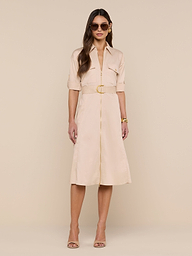 A woman wears the Callen Shirt Dress—a belted, light beige, knee-length shirt dress with short sleeves and button detailing—styled with sunglasses, nude heels, and a gold bracelet as she stands against a plain white background.