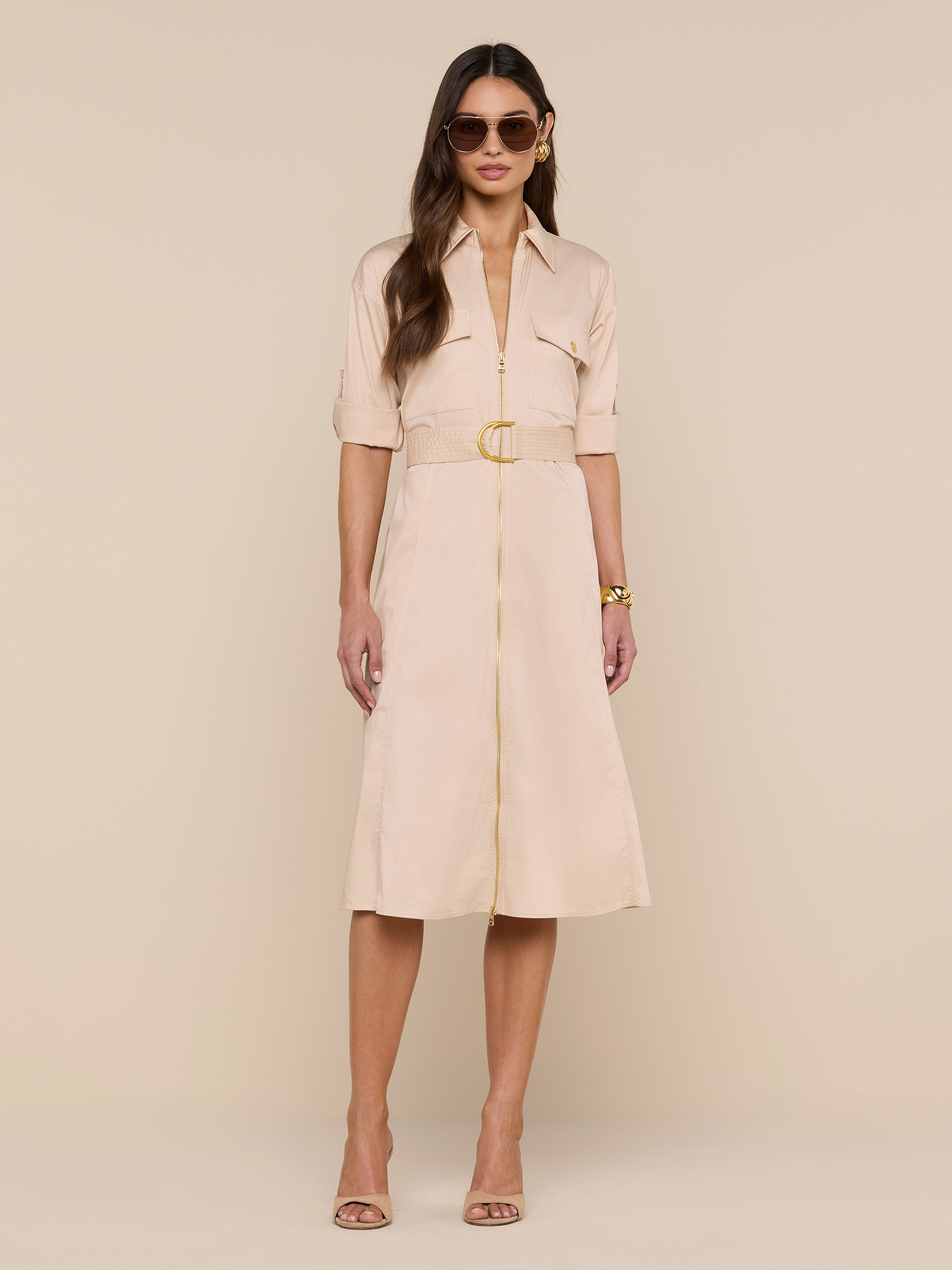 A woman wears the Callen Shirt Dress—a belted, light beige, knee-length shirt dress with short sleeves and button detailing—styled with sunglasses, nude heels, and a gold bracelet as she stands against a plain white background.