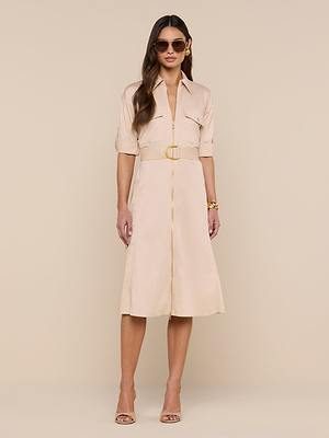A woman poses against a beige backdrop wearing the Callen Shirt Dress—a light beige, collared stretch cotton-blend dress with rolled sleeves and a belt—styled with neutral heels, sunglasses, and gold jewelry.