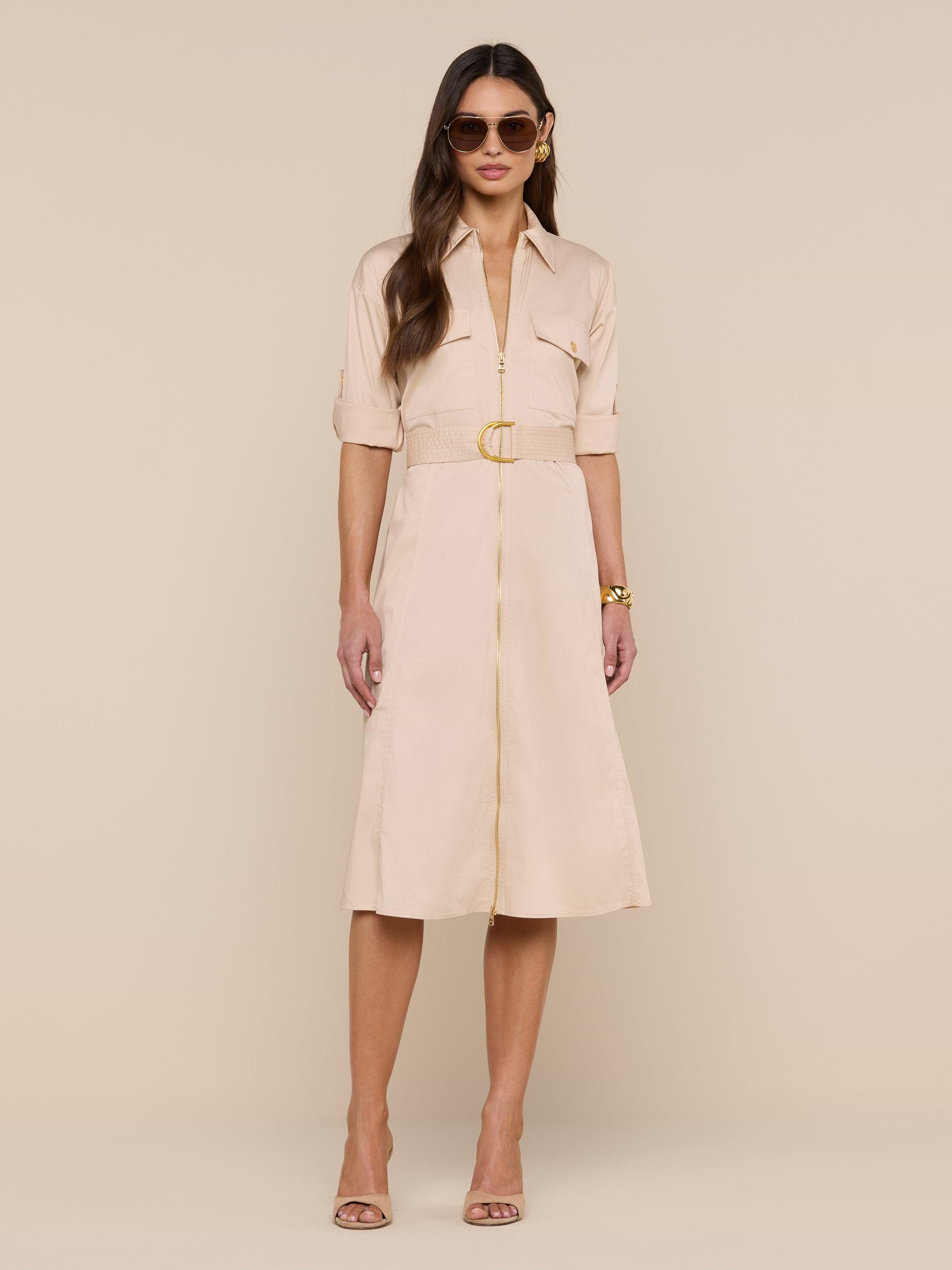 A woman wears the Callen Shirt Dress—a belted, light beige, knee-length shirt dress with short sleeves and button detailing—styled with sunglasses, nude heels, and a gold bracelet as she stands against a plain white background.