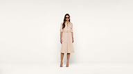 Woman wearing sunglasses, a beige belted midi dress, and matching sandals stands against a plain white background.