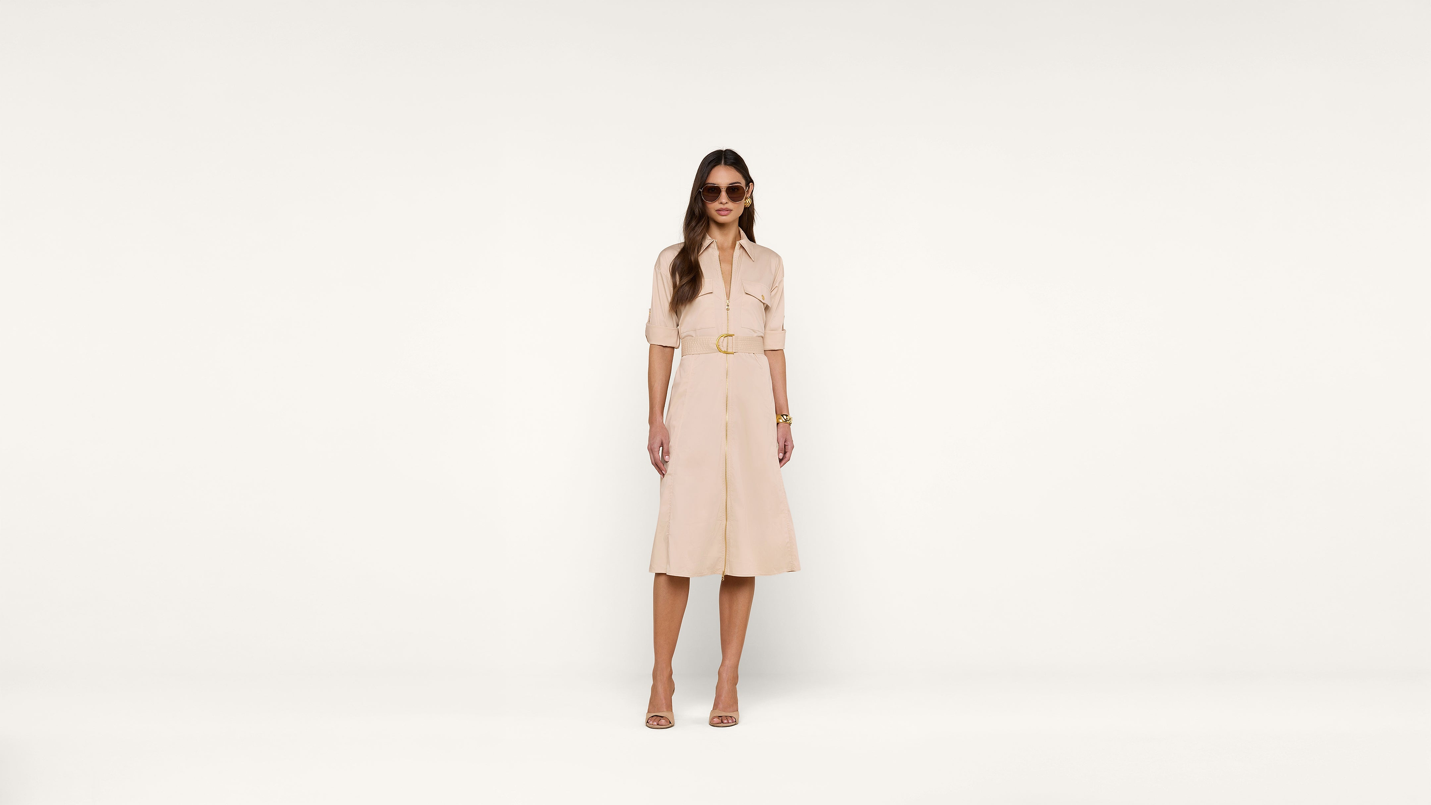 Woman wearing sunglasses, a beige belted midi dress, and matching sandals stands against a plain white background.