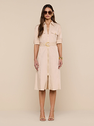 A woman wearing sunglasses and the Callen Shirt Dress—a light beige, belted design—stands against a plain white background, styled with neutral high-heeled sandals and gold bracelets.