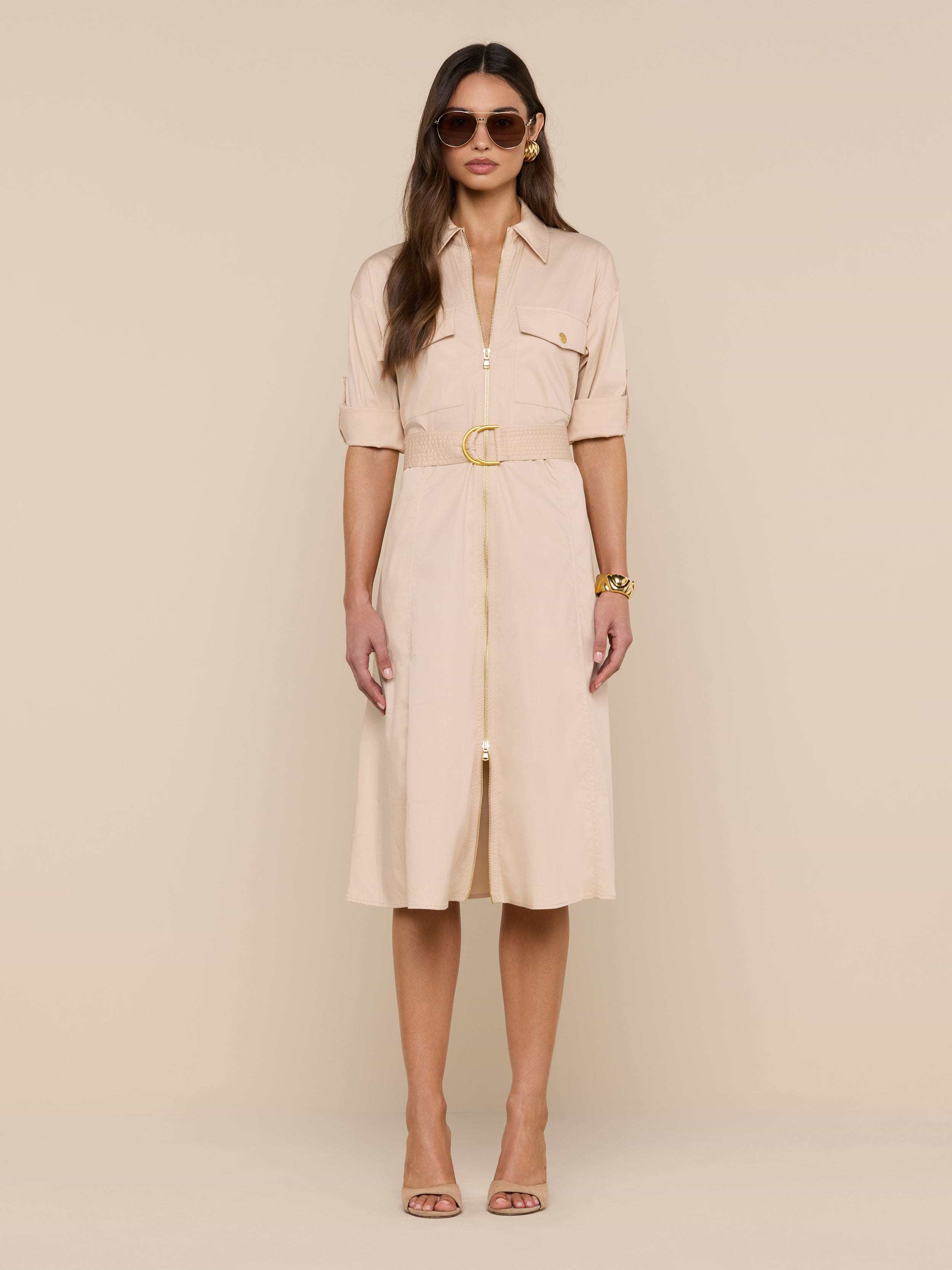 A woman wearing sunglasses and the Callen Shirt Dress—a light beige, belted design—stands against a plain white background, styled with neutral high-heeled sandals and gold bracelets.