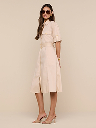 A woman wearing sunglasses, the Callen Shirt Dress with rolled sleeves and a belted waist in beige, paired with nude heels, stands against a plain white background, looking confidently at the camera.