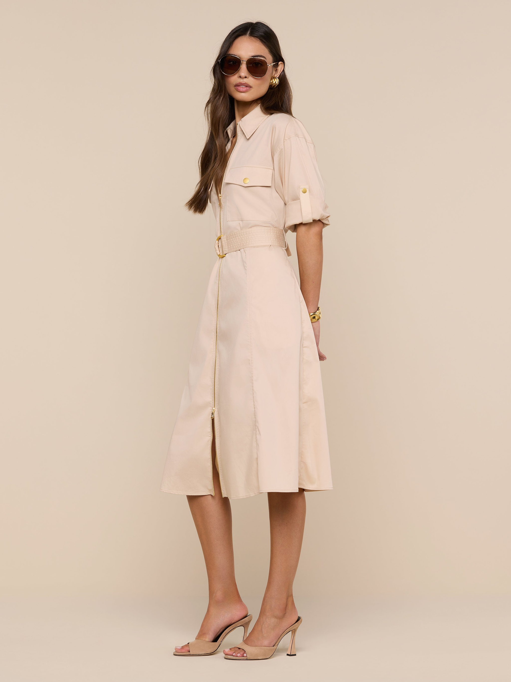 A woman wearing sunglasses, the Callen Shirt Dress with rolled sleeves and a belted waist in beige, paired with nude heels, stands against a plain white background, looking confidently at the camera.