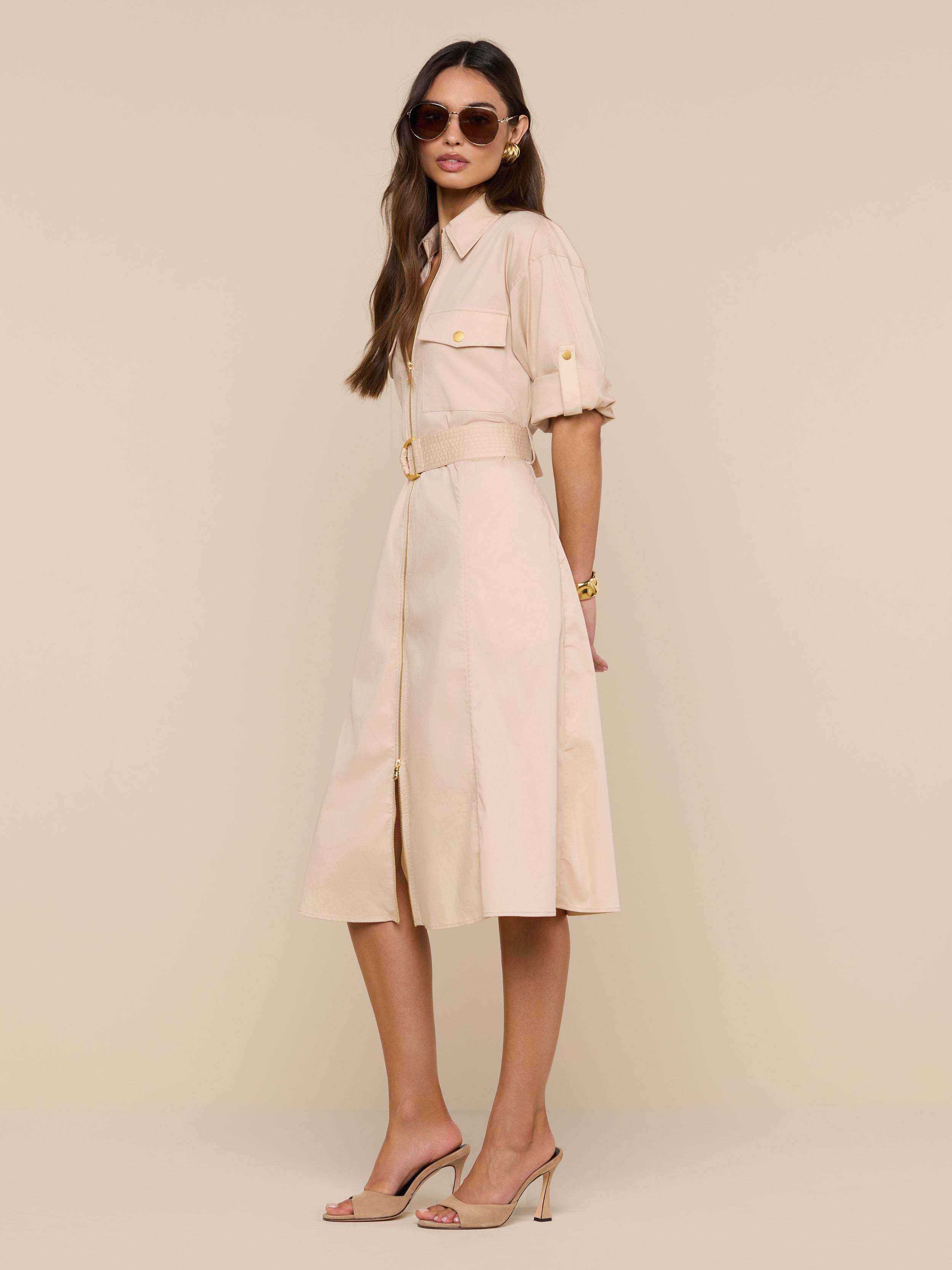 A woman wearing sunglasses, the Callen Shirt Dress with rolled sleeves and a belted waist in beige, paired with nude heels, stands against a plain white background, looking confidently at the camera.