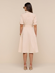 A woman stands facing away, wearing the Callen Shirt Dress—a knee-length, light beige belted dress with short sleeves—paired with nude high heels against a plain white background.