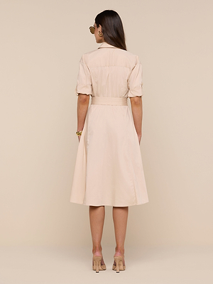 A woman with long brown hair stands facing away, wearing the Callen Shirt Dress—a light beige, knee-length stretch cotton-blend piece with short sleeves and a fitted waist. She wears sunglasses and heels, set against a plain beige background.