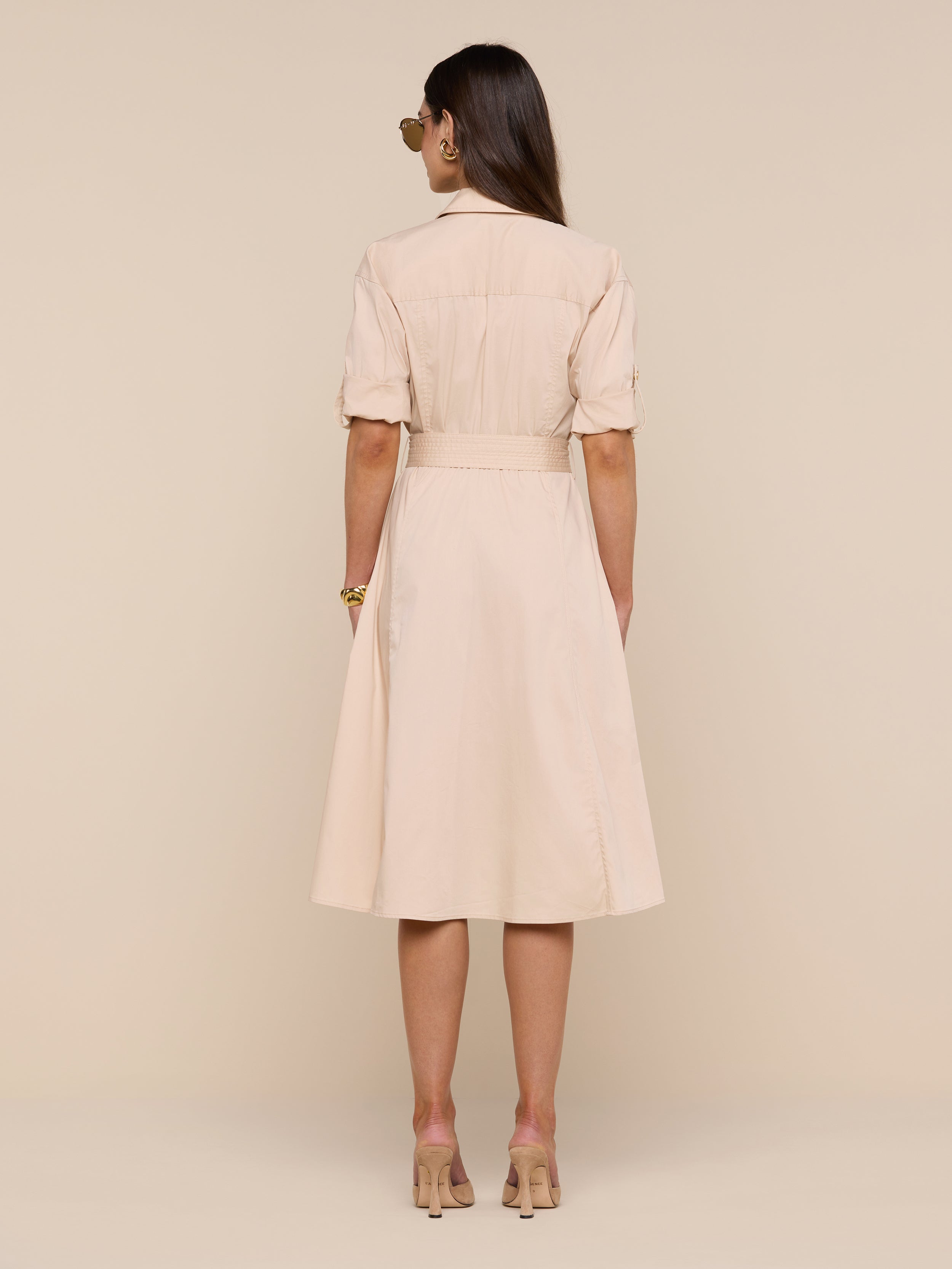 A woman stands facing away, wearing the Callen Shirt Dress—a knee-length, light beige belted dress with short sleeves—paired with nude high heels against a plain white background.