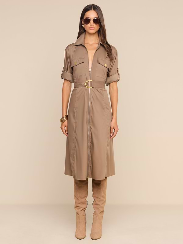 Une femme aux cheveux longs, arborant des bracelets en or à un poignet, porte la robe-chemise Callen — confectionnée dans une popeline douce en mélange de coton de couleur beige clair, dotée dune fermeture éclair apparente sur le devant, dune ceinture à la taille et de manches retroussées. Elle lassocie à des bottes en daim montant jusquaux genoux et à des lunettes de soleil, posant devant un fond beige uni.