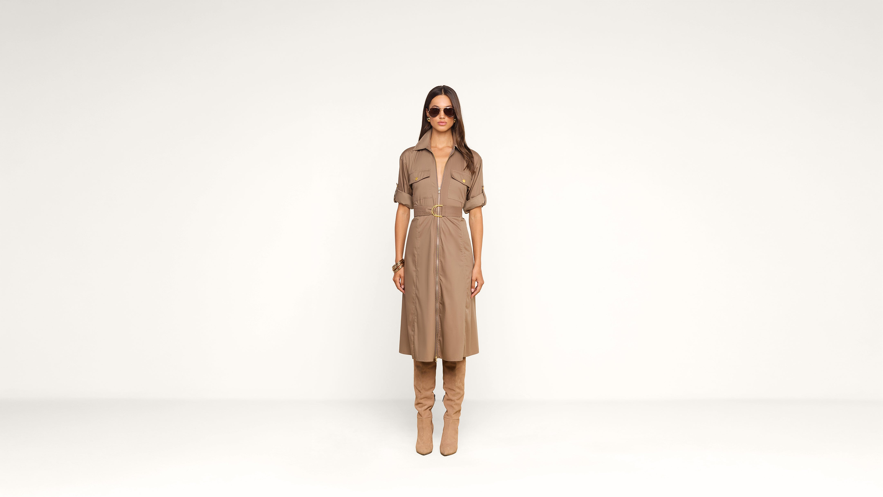 A woman wearing sunglasses, a tan belted dress, and matching knee-high boots stands against a plain white background, facing forward.