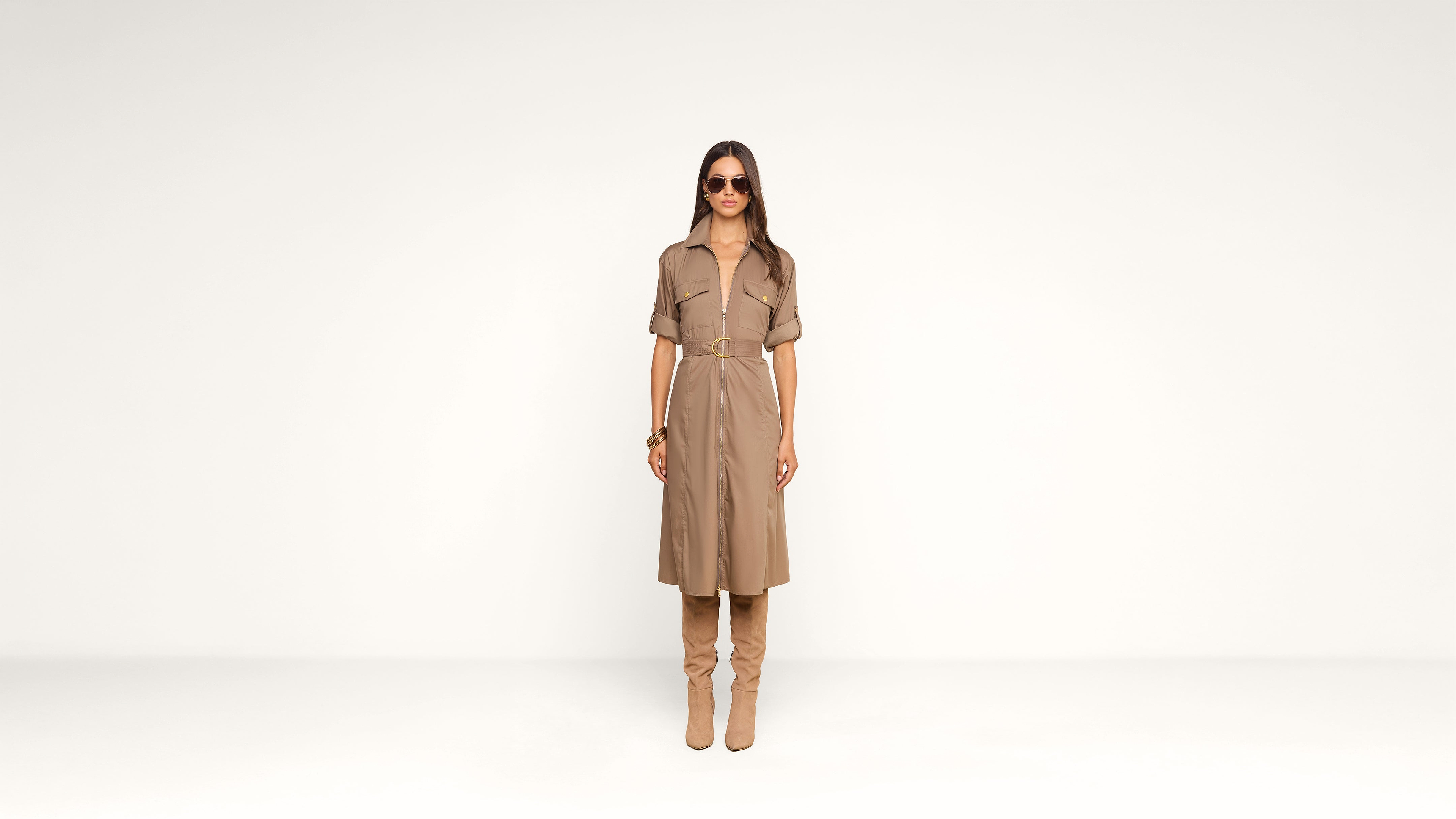 A woman wearing sunglasses, a tan belted dress, and matching knee-high boots stands against a plain white background, facing forward.